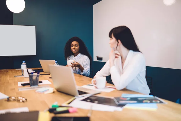 若い多民族の女性の同僚は カジュアルな服を着てコーヒーを飲みながら オフィスでノートパソコンや文書と机の上に座って話しています — ストック写真
