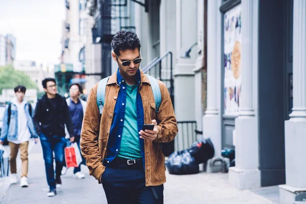 Handsome focused interested stylish male in casual outfit browsing and texting with smartphone while looking down and walking along street with people on urban background