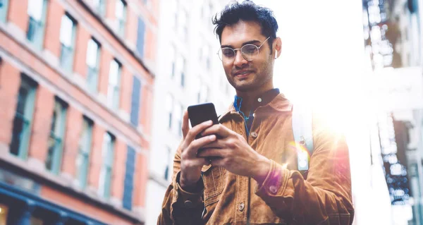 携帯電話で茶色のスエードジャケットメッセージを身に着けている眼鏡でハンサムなUnshavenヒスパニック男性からと晴れた日にニューヨークの街の歩道に立っている間笑顔 — ストック写真