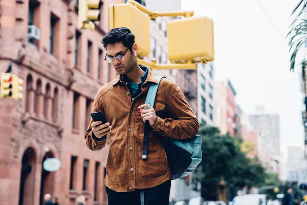 真剣にハンサムな民族カジュアル大人男性で眼鏡でバックパックタイプスマートフォンで忙しい街の路上で日光の中に立つ — ストック写真