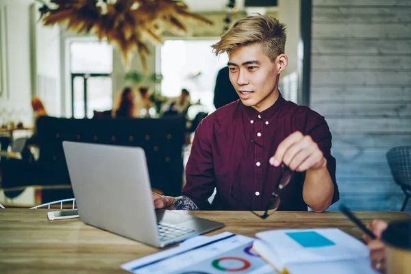 Odaklanmış ciddi Asyalı erkek, sarı saçlı, günlük eflatun gömlekli, gözlük takan, yaratıcı iş yerinde dizüstü bilgisayar kullanan, arka planda meslektaşları olan.
