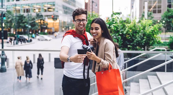 带着相机和购物袋的多种族快乐男女站在城市背景下 看着印好的照片笑着 — 图库照片