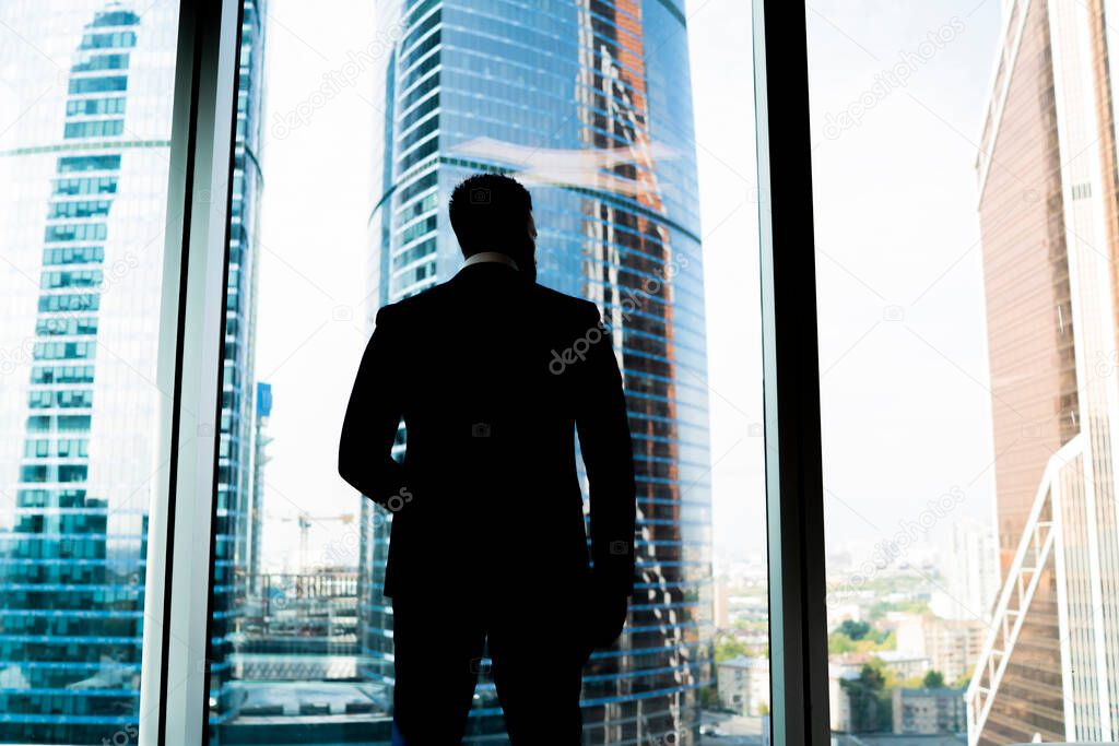From below back view of faceless confident man in suit standing in darkness against office window looking away on skyscrapers