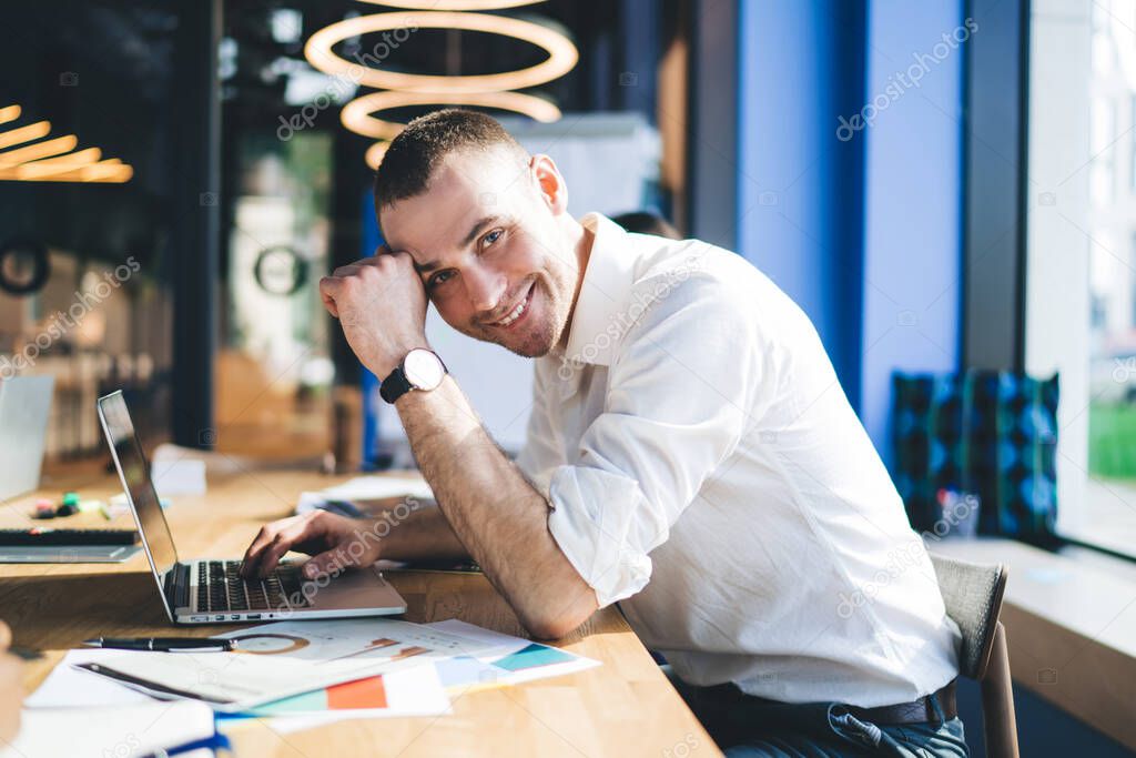 Content man sitting at desk in contemporary office while using laptop and studying documents while looking at camera on blurred background