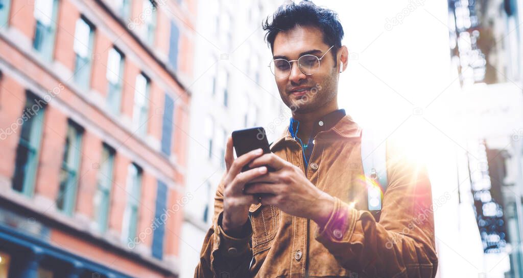 From below handsome unshaven Hispanic male in eyeglasses wearing brown suede jacket messaging on cellphone and smiling while standing on New York city street sidewalk on sunny day