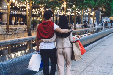 Günümüz modasına uygun giyinen, sevgiyle sarılan ve alışveriş torbalarıyla şehir caddesinde yürüyen çiftin arka planı.