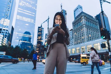 Mutlu, etnik ve modern bir bayanın resmi kıyafetleri içinde şehir caddesinde gökdelenlerin önünde zevkle akıllı telefonlara bakmasından.