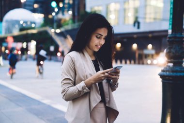 Düşünceli, modern stil sahibi, bej takım elbiseli, akıllı telefon kullanan, gökdelenleri ve ışıkları olan bir kadın. 