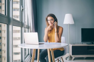 Sarı kolsuz bluzuyla beyaz yuvarlak masada oturan, evinde kameraya bakarak akıllı telefondan iletişim kuran, memnun genç bir kadın.