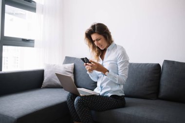 Oturma odasındaki kırışık suratlı dizüstü bilgisayarla koltukta oturmuş kafası karışık bir şekilde cep telefonu kullanan genç bir kadın.