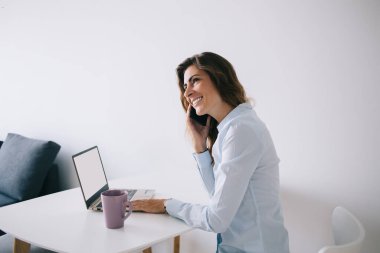 Bardak ve dizüstü bilgisayarın yanında oturan beyaz masada boş yeri olan ve telefonda gülümseyen genç bir kadının yan görüntüsü.