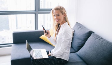 Beyaz ofis gömlekli, rahat koltukta oturan, dizüstü bilgisayarlı, akıllı telefon kullanan ve kameraya bakan serbest kadın.