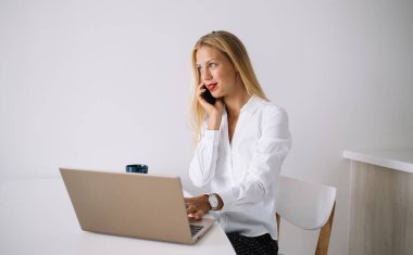 Konsantre olmuş genç sarışın kadın beyaz masada mavi bardaklı bluzuyla oturuyor akıllı telefonuyla konuşuyor evde dizüstü bilgisayarını kullanıyor ve gözünü kaçırıyor.