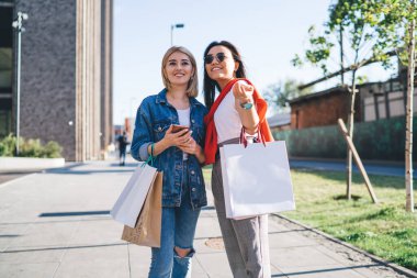 Neşeli kadın kamerayı işaret ediyor. Güneşli bir günde kot pantolon giymiş, alışveriş torbalarıyla rahatlıyor.