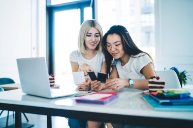 Modern neşeli kız arkadaşlar çalışma odasında masalarında oturup projeye hazırlanırken anlık fotoğraflara ve yapışkan notlara bakarlar.
