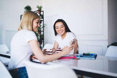Doğal ışıklandırılmış çağdaş kafe masasında otururken gülümseyen kız arkadaşıyla cep telefonunu paylaşan sıradan giyinmiş bir arkadaşın yan görüntüsü. 