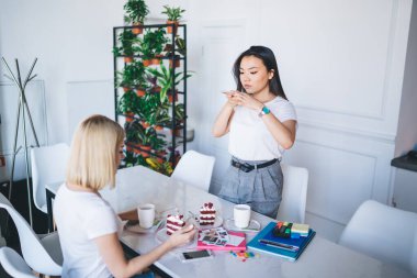 Beyaz tişörtlü çok ırklı meslektaşlar, ellerinde kek fincanları, kahve fincanları, beyaz modern ofis alanındaki işaretler ve arka plandaki saksılarda bitkilerle masada oturuyorlar.  