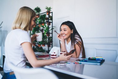 Beyaz tişörtlü çok ırklı kadınlar cep telefonlarıyla mesajlaşıyorlar. Masalarında kahve ve kekle modern beyaz stüdyoda oturuyorlar.