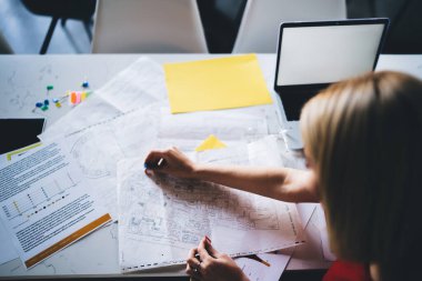 Yukarıdan gelen konsantre kadın belgelerle çalışıyor ve kağıt okuyor. Masada önemli noktalara dizüstü bilgisayar ve sarı kağıt yapıştırıyor. 