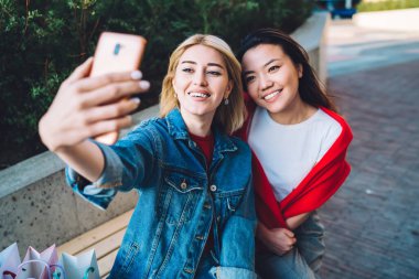 Yukarıdan gelen mutlu, çekici, çeşitli bayan arkadaşlar akıllı telefondan selfie çekiyorlar. Şehir sokağındaki bankta birlikte dinleniyorlar.