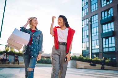 Sıradan giyinmiş neşeli bir kadın, telefonu elinde, kot elbiseli, alışveriş çantalı, gülümseyen bir kadınla modern şehrin sokaklarında birbirine bakıyor.