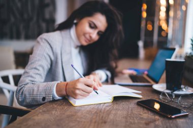 Uzun siyah saçlı, günlük giysiler içinde genç ve zeki bir bayan kafeteryada dizüstü bilgisayarla masaya oturmuş not defteri tutuyor.  