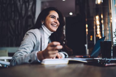 Kahkahalarla gülen, uzun siyah saçlı, günlük kıyafetlerle neşeli, kafede dizüstü bilgisayarla oturup notlar alan bir kadın.  