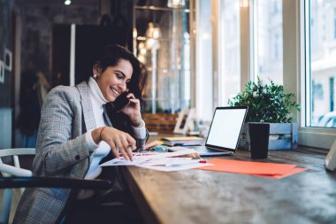 Mutlu bayanın iş arkadaşıyla telefonda konuşurken bilgisayarla kafede oturarak evrakları analiz ederken yan görüntüsü.