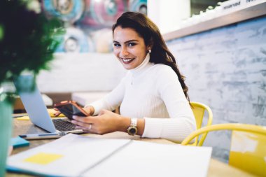 Kafeteryada oturmuş kameraya bakarken dizüstü bilgisayar ve cep telefonu kullanarak projede çalışan neşeli, genç esmer kadın.