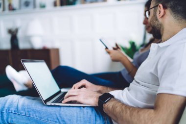 Yandan bakıldığında, gözlüklü, sakalsız, yetişkin bir erkeğin laptopta oturup tabletli bir kadına bulanık arka planda baktığı görülüyor.