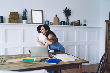 Beyaz tişörtlü bir adam ve dişlek gülüşlü, genç, siyah saçlı bir kadın modern iş yerinde birbirini kucaklıyor ve tebrik ediyor.