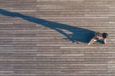 Yoga kıyafetleri içindeki sağlıklı bir kadın ahşap zeminde, uzun gölgesi düşerken poz veriyor.