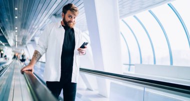 Modern cep telefonu zımbırtısıyla çevrimiçi sohbet etmek için boş zamanlarında elektrikli merdivenlerde dikilip kameraya bakan, şık giyinmiş, modern çağdan kalma hippi bir adamın yarım boy portresi.