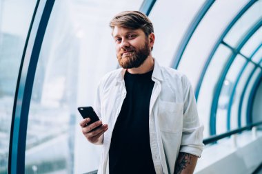 Yakışıklı hipster blog yazarının yarım boy portresi, günlük beyaz gömlekli cep telefonu cihazını elinde tutuyor ve boş zamanlarında kameraya bakıyor, teknoloji ve iletişim kavramı.