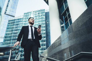 Elinde cep telefonu olan klas siyah takım elbiseli, sakallı genç bir yöneticinin modern şehirde merdivenlerden gökdelenlerle inmesinden.