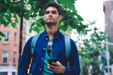 Kot gömlekli, sırt çantalı, cep telefonlu sıradan modern bir adam New York caddesinde duruyor ve başka tarafa bakıyor.