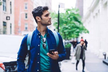Sırt çantalı, modaya uygun mavi tişörtlü genç ve yakışıklı bir adam cep telefonu tutuyor ve New York sokaklarına bakıyor.