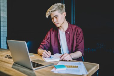 Beyaz tişörtlü, sarı saçlı ve kırmızı tişörtlü konsantre Asyalı bir erkek masada belgelerle oturuyor.