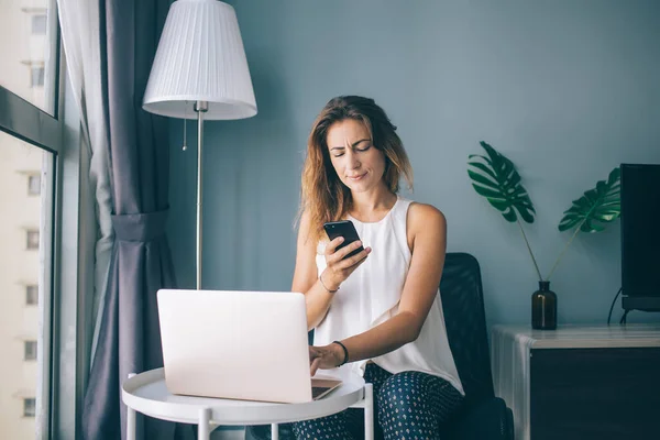 Focused serious young woman in white sleeveless blouse sitting at round table working on laptop and typing on smartphone at home