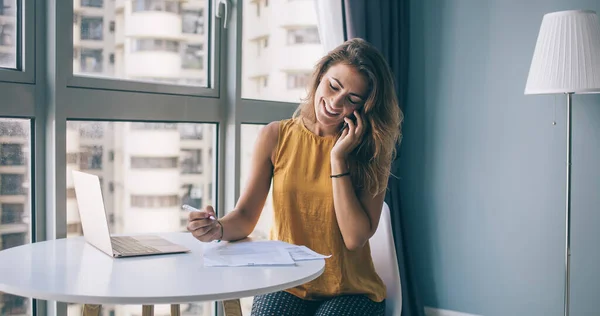 Smiling young female with eyes closed and pen in casual clothes sitting at desk with papers and laptop while talking on smartphone at home