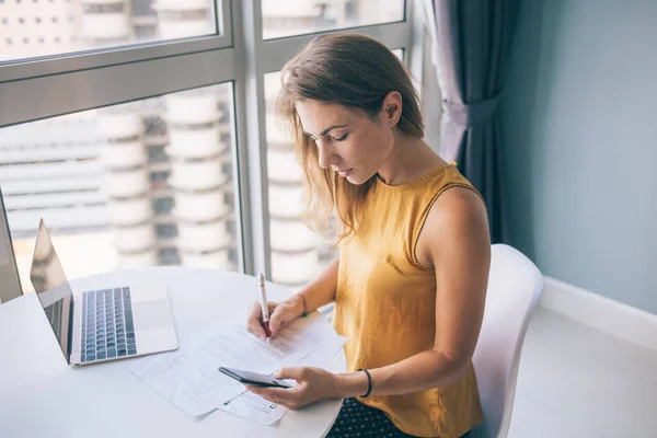 From above serious young woman in yellow sleeveless blouse sitting at white round table with laptop and paper typing on smartphone at home