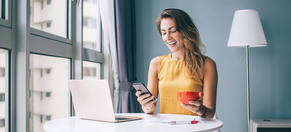 Excited woman enjoying coffee time for online chatting with virtual friends via modern cellphone gadget, positive Caucasian female blogger reading funny comments in social network during distance job