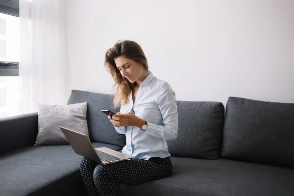 Modern adult woman in shirt texting on phone while sitting on sofa with laptop on knees  in living room of apartment