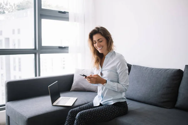 Young business lady in shirt and trousers chatting on phone sitting on sofa with computer nearby in living room at apartments smiling