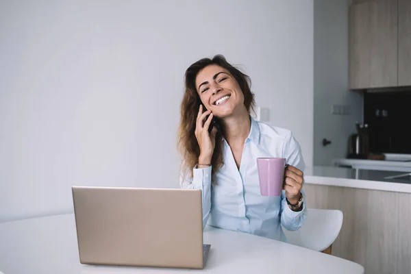身穿白衬衫 快乐的成年女性坐在桌旁 一边听着笔记本电脑一边喝咖啡 一边闭着眼睛微笑 — 图库照片