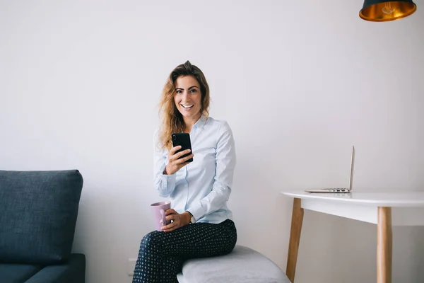 Młoda Pracownica Siedzi Skrzyżowanymi Nogami Smartfon Kubek Napojem Patrząc Kamerę — Zdjęcie stockowe