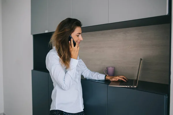 穿着蓝色工作服 面带微笑的职业女性 一边站在家里一边用笔记本电脑打字 一边用智能手机友好地交谈 — 图库照片