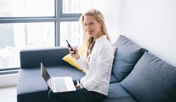 身穿白衬衫的女自由撰稿人坐在舒适的沙发上 膝上型笔记本电脑 一边用智能手机一边看相机 一边看侧面 — 图库照片