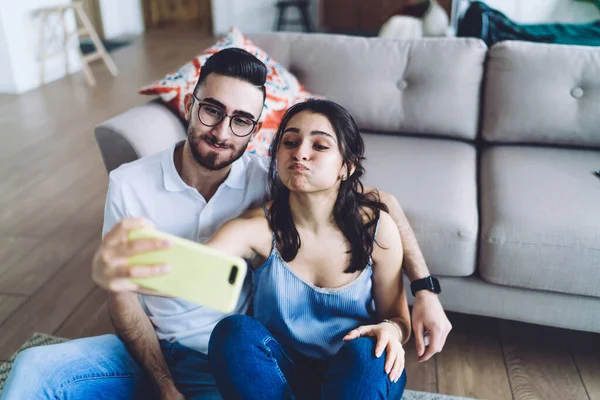 Happy sweet couple in comfy clothes relaxing and making faces on floor by couch while shooting selfie and using laptop in flat on blurred background