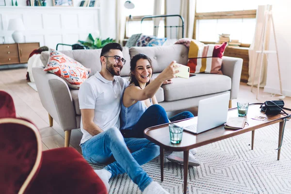 Happy sweet couple in casual wear relaxing and cuddling by modern couch while shooting selfie and using laptop in apartment with contemporary interior